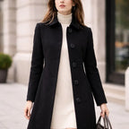 Woman wearing a black long coat with large buttons over a cream turtleneck dress outdoors.
