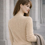 Woman in a beige ribbed turtleneck sweater holding a gray coat, standing outdoors.